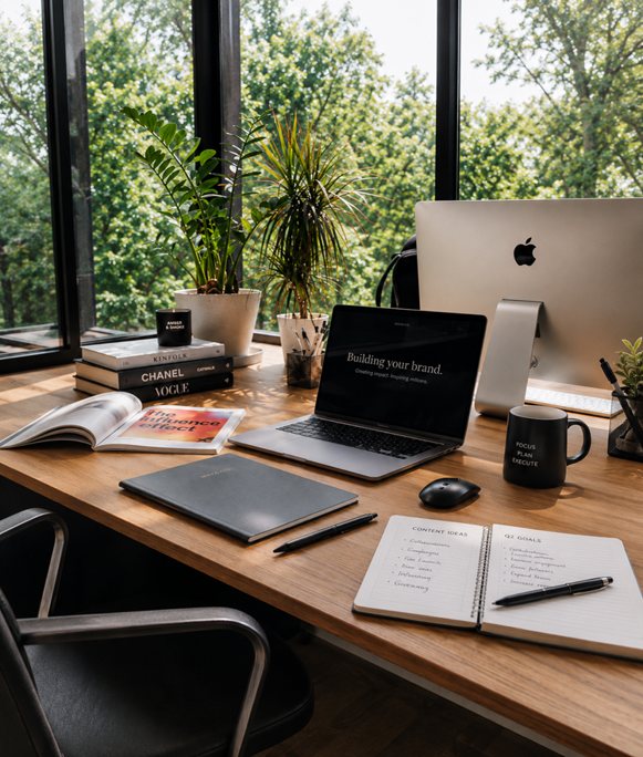 A modern desk with an iMac and laptop beside plants and large windows